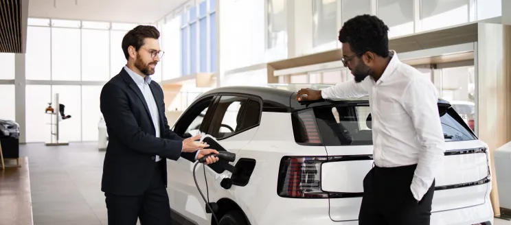 Dealer sells electric SUV to businessman in car dealership. They discuss vehicle features. Business environment and modern electric vehicle concept.