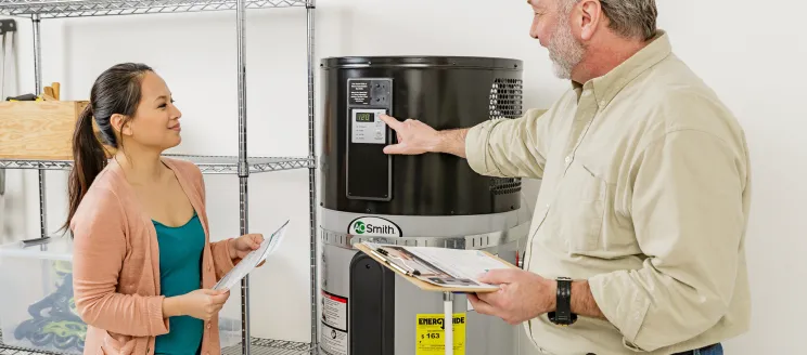 A contractor explains the settings on a heat pump water heater to a homeowner, who is holding informational paperwork. 
