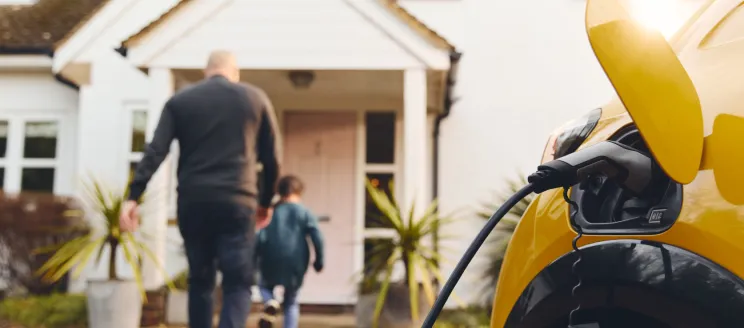 Stock image of an electric vehicle charging as family walks into home in the background