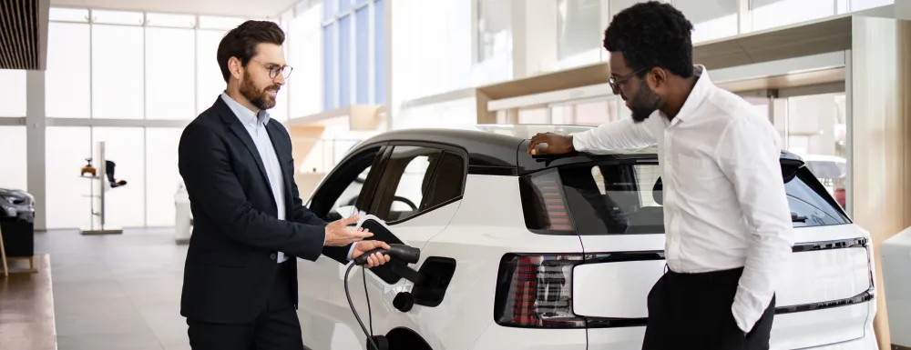 Dealer sells electric SUV to businessman in car dealership. They discuss vehicle features. Business environment and modern electric vehicle concept.