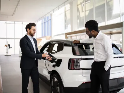Dealer sells electric SUV to businessman in car dealership. They discuss vehicle features. Business environment and modern electric vehicle concept.